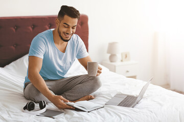 Smiling man sits cross-legged on a bed while holding a coffee cup. He looks at a notebook next to his laptop in a bright, cozy bedroom, enjoying his morning routine.