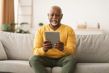 A senior man sits comfortably on a gray couch in a well-lit living room. He holds a tablet and smiles, enjoying his time while engaging with technology. The atmosphere is warm and inviting.