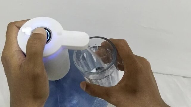 view of a man's hand carrying a glass filled with mineral water to drink from a gallon using an electric gallon pump
