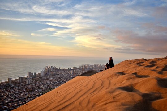 Mujer joven sentada en la duna Cerro Drag&oacute;n con la ciudad de Iquique y el oc&eacute;ano pac&iacute;fico al fondo, en el norte de Chile.
