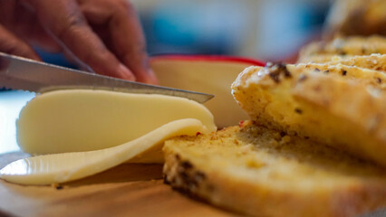 Close-up of a hand slicing cheese onto warm artisan bread. Natural light, food preparation scene ideal for cooking, bakery, recipe, and culinary content.
