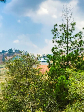 Mountain landscape with lush green trees, blue sky, and clouds, viewed from a hillside road in Indonesia, showing natural scenery and peaceful outdoor atmosphere.
