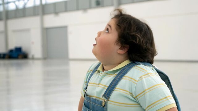 Chubby boy child kid backpack in denim overall and striped polo surprised looking up with open mouth candid expression in indoor hangar setting