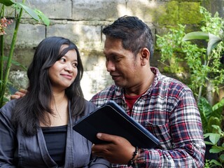 Happy couple sharing a moment while viewing a digital tablet together in a lush, green outdoor garden. Great for relationship, technology, lifestyle, and leisure concepts.