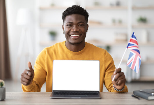Positive african american man sitting at table at home, showing flag of UK and laptop with blank screen, mockup, copy space. Charismatic black guy studying English on Internet, having online course