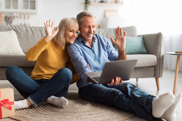 An elderly couple sits together on the floor, smiling and waving at a laptop. They are in a cozy living room, showcasing warmth and connection through technology during a sunny afternoon.