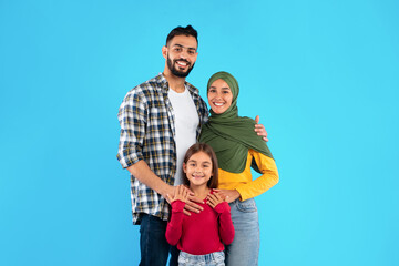 A happy family stands together with the father wearing a plaid shirt, the mother in a green hijab and yellow top, and their daughter in a red shirt. They all show joy and love in this bright setting.