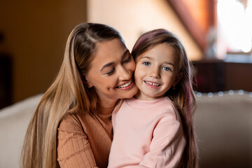 A mother and her young daughter smile and hug each other in a cozy living room. The warm sunlight filters through the window, creating a joyful and loving atmosphere.
