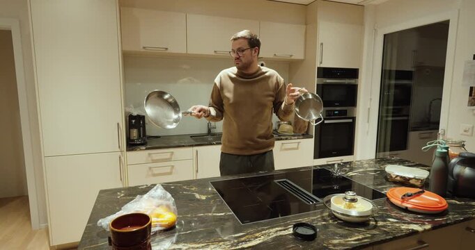 A man stands clueless in a modern kitchen, holding two frying pans with a confused expression. He appears overwhelmed and unsure about cooking, not knowing what to do next.