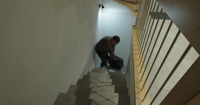 A man lifts and carries a heavy suitcase up a staircase, showing physical effort, travel preparation, exhaustion, and the challenge of moving luggage through narrow stairways.