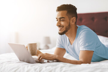 A young man is lying on a bed, smiling while typing on his laptop. He holds a cup of coffee, enjoying a relaxed morning in a bright and comfortable bedroom setting.