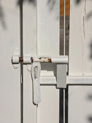 Close-up detail of a white wooden gate secured by a rustic metal slide latch, illuminated by bright sunlight with contrasting shadows.