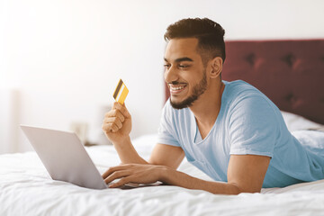 A smiling young man lays on his bed, using a laptop to shop online. He holds a credit card in one hand, enjoying a comfortable moment. The room is bright and cozy, creating a relaxed atmosphere.