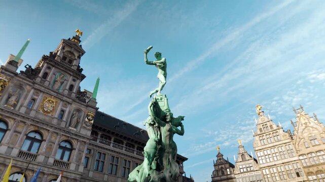 Low-angle view of the iconic Brabo Fountain statue in the Grote Markt of Antwerp, Belgium. Historic guildhalls and the City Hall frame the legendary bronze sculpture under a blue sky