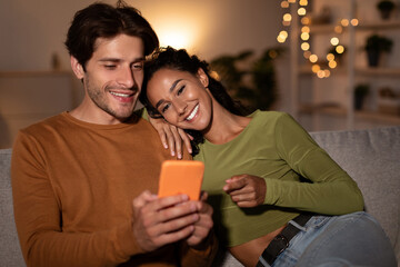 Couple sitting on a comfortable sofa at home is happily using a smartphone. The woman points at the screen, smiling at her husband. The scene is cozy with warm lighting in the living room.
