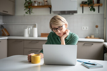 Thoughtful middle aged businesswoman enjoying morning coffee and reading work documents on laptop, looking at screen, resting head on hand. Pensive lazy business lady works on computer in kitchen.