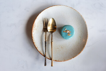 Premium French dessert concept. Single turquoise macaron decorated with edible gold flakes, placed beside brass spoon and fork. Minimalist flat lay food photography.