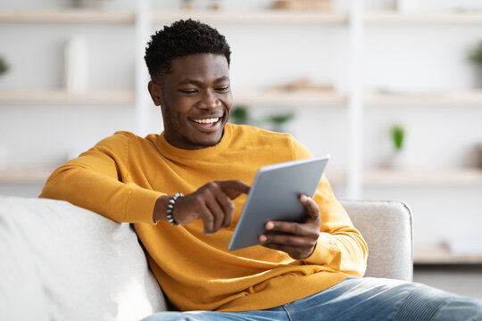 Smiling african american young man sitting on couch with digital tablet, chatting with ladies, using dating mobile application on modern pad, home interior, copy space. Gadgets addiction concept - Powered by Adobe