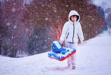 Happy Boy Walking Up Snowy Hill with Inflatable Sled &ndash; Winter Adventure in Falling Snow. Fun Seasonal Ride