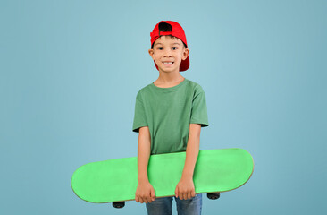 A child stands smiling while holding a green skateboard with both hands. He wears a green t-shirt and a red cap, ready to enjoy skating indoors or outdoors.