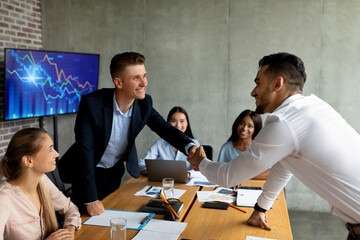 Two men shake hands after closing a deal during a corporate meeting in a modern office. The atmosphere is positive, with smiling faces and colleagues observing the agreement.