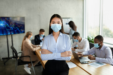 Confident young asian businesswoman wearing a medical mask stands with folded arms in an office. Colleagues are engaged in discussion and working at a conference table during a corporate meeting.