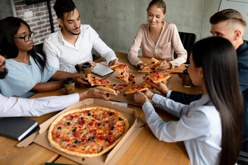 Colleagues from diverse backgrounds share a joyful lunch break in the conference room, enjoying pizza and laughing together while seated around a table filled with food.