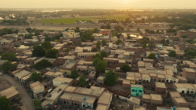 Forward drone glide over Sher Shah City village in Multan Punjab capturing the golden hazy sunlight bathing the rural houses and green fields at sunset.