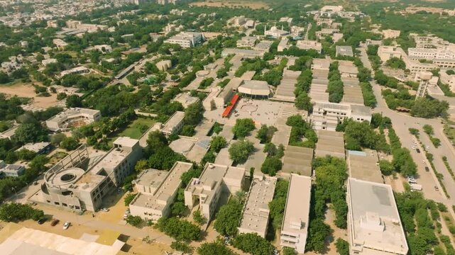 Drone descending and orbiting with a downward tilt, showcasing the green vegetation, trees, and architecture of NED University campus during an intense sunny day.