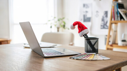 Creative workspace with a tablet, stylus, color swatches, and laptop with Christmas hat on pencil holder and colorful stationery