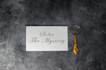 A white card displaying the phrase “Solve the Mystery” stands on a textured dark surface, accompanied by an elegant gold magnifying glass, creating a detective-inspired and suspenseful visual theme