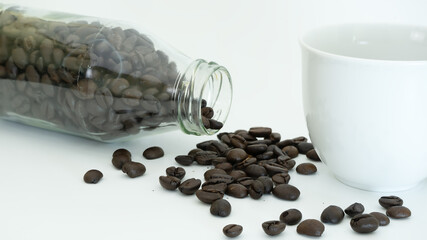 Spilled roasted coffee beans from glass bottle on white background with white cup.