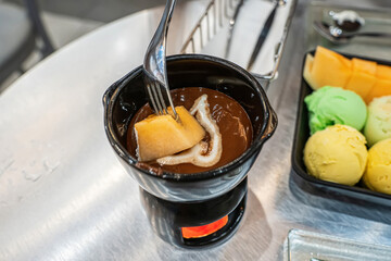 A fork dipping a piece of yellow melon into a pot of warm chocolate fondue sauce.
