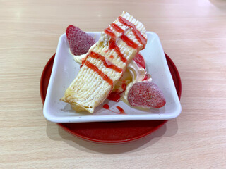 A slice of creamy mille crepe cake topped with sweet strawberry sauce and fresh strawberries, served on a white plate, ready to eat as a delicious dessert.
