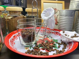 A vintage-style coffee preparation setup featuring roasted coffee beans on a tray, traditional fabric filters, glasses, and storage jars on a wooden shelf.