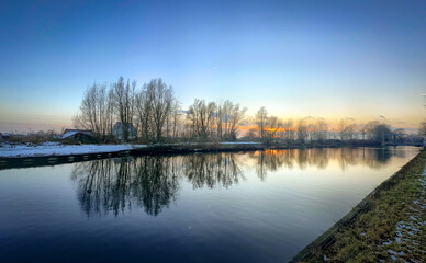 A tranquil winter sunset casts vibrant reflections on the river, with silhouetted trees along the banks