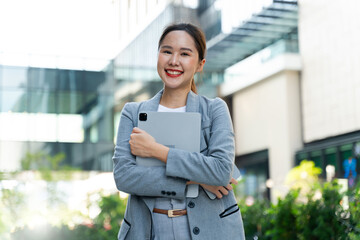 Confident young Asian businesswoman smiling and standing outside a modern office building. A happy professional woman using her smartphone in the city, showcasing urban lifestyle and technology.