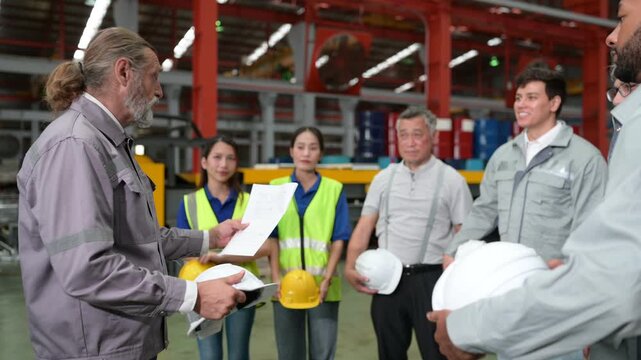 Diverse group of factory workers and engineers attending a safety meeting. A senior manager leads the morning briefing with staff wearing hard hats and vests in an industrial manufacturing plant.