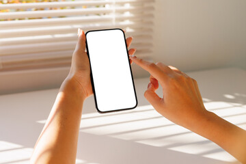 Woman's hand holding a smartphone with an empty screen, tapping it, with sunlight and blinds in the background. Technology concept