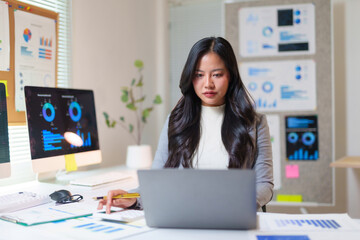 Asian businesswoman working with laptop and computer screens displaying charts, focused on data analysis and business growth strategy
