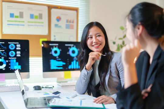 Two smiling Asian businesswomen discussing business strategy and data analysis during a meeting in a contemporary office setting - Powered by Adobe