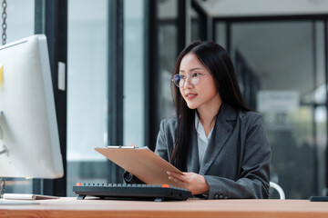 Young Asian woman wearing glasses, holding a clipboard and looking at a computer monitor in a modern corporate office