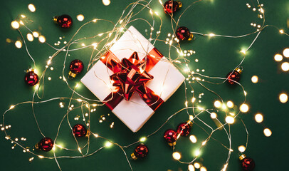 Christmas gift box with red decorations, Christmas light garland, bokeh, glitter on green holiday background. Merry Christmas and Happy New Year greeting card. Xmas and winter background, top view