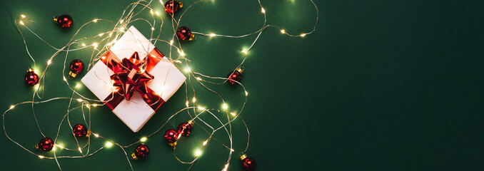 Christmas gift box with red decorations, Christmas light garland, bokeh, glitter on green holiday background. Merry Christmas and Happy New Year greeting card. Xmas and winter background, top view