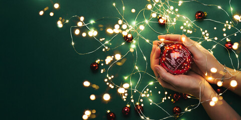 Female hands holding red Christmas ornament on green background with Christmas light garland, bokeh,glitter. Merry Christmas and Happy New Year greeting card. Xmas and winter holidays, top view