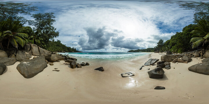 A peaceful sandy beach with large boulders, gentle turquoise waves, and lush green vegetation beneath a cloudy sky. Anse Intendance, Seychelles. 360VR.