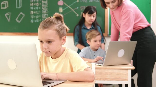 Caucasian boy focus on programing system while looking at camera surrounded by diverse student coding engineering prompt. Happy teacher support children working on laptop at computer lesson. Pedagogy.