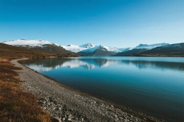Snow-capped mountains reflected in a calm lake surrounded by rocky shoreline and autumnal vegetation under a clear blue sky