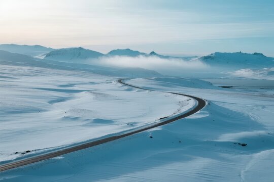 A winding road cuts through a vast snowy landscape with distant mountains and mist under a clear sky - Powered by Adobe
