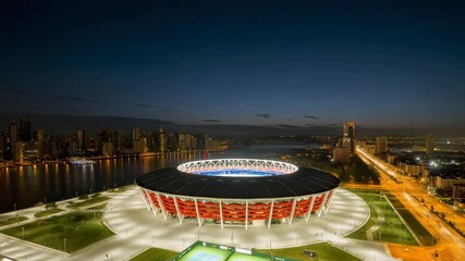 Panoramic aerial view of a brightly illuminated modern sports stadium complex situated beside a major city skyline at twilight near a large body of water - Powered by Adobe